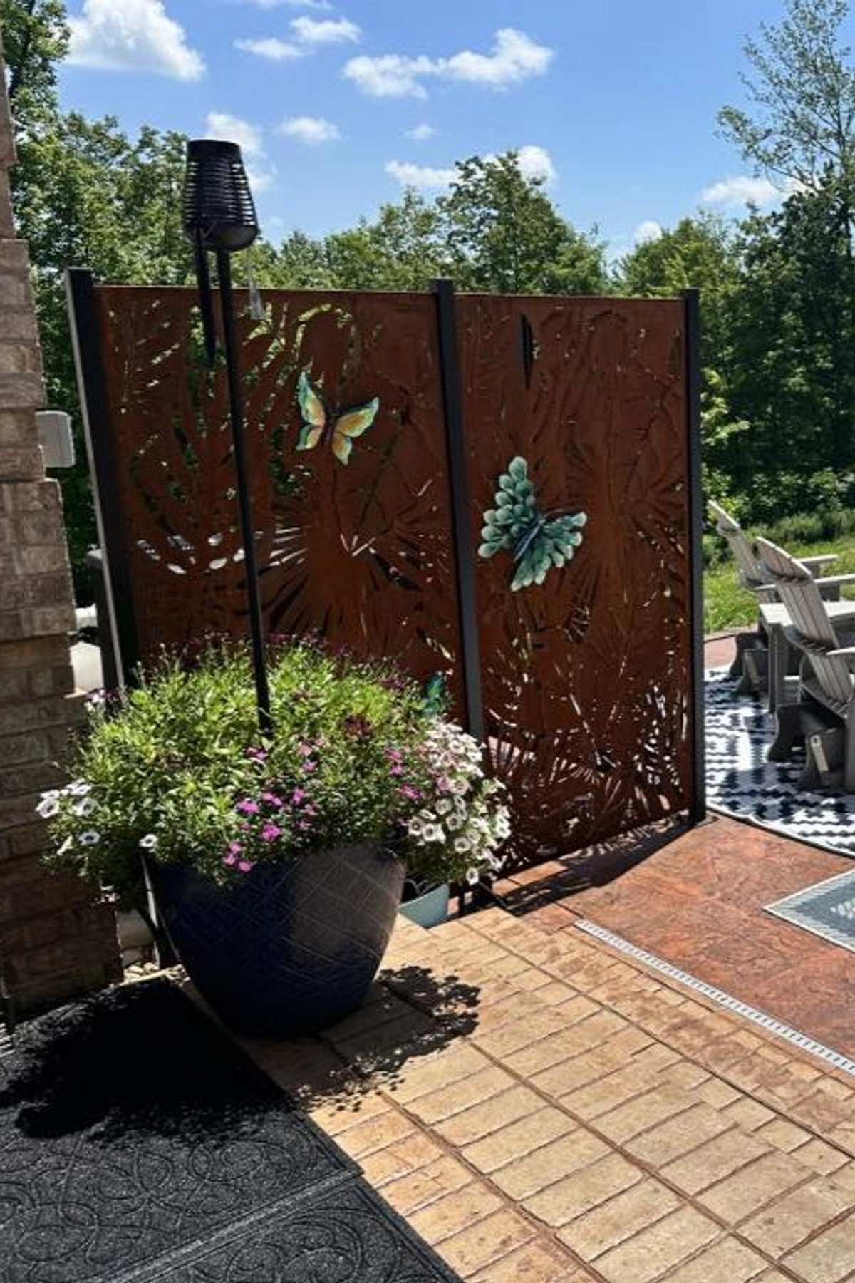 Backyard privacy screen made of corten steel with decorative cutouts and featuring a butterfly accents, placed beside a large planter filled with summer flowers, patio chairs and greenery in the background.