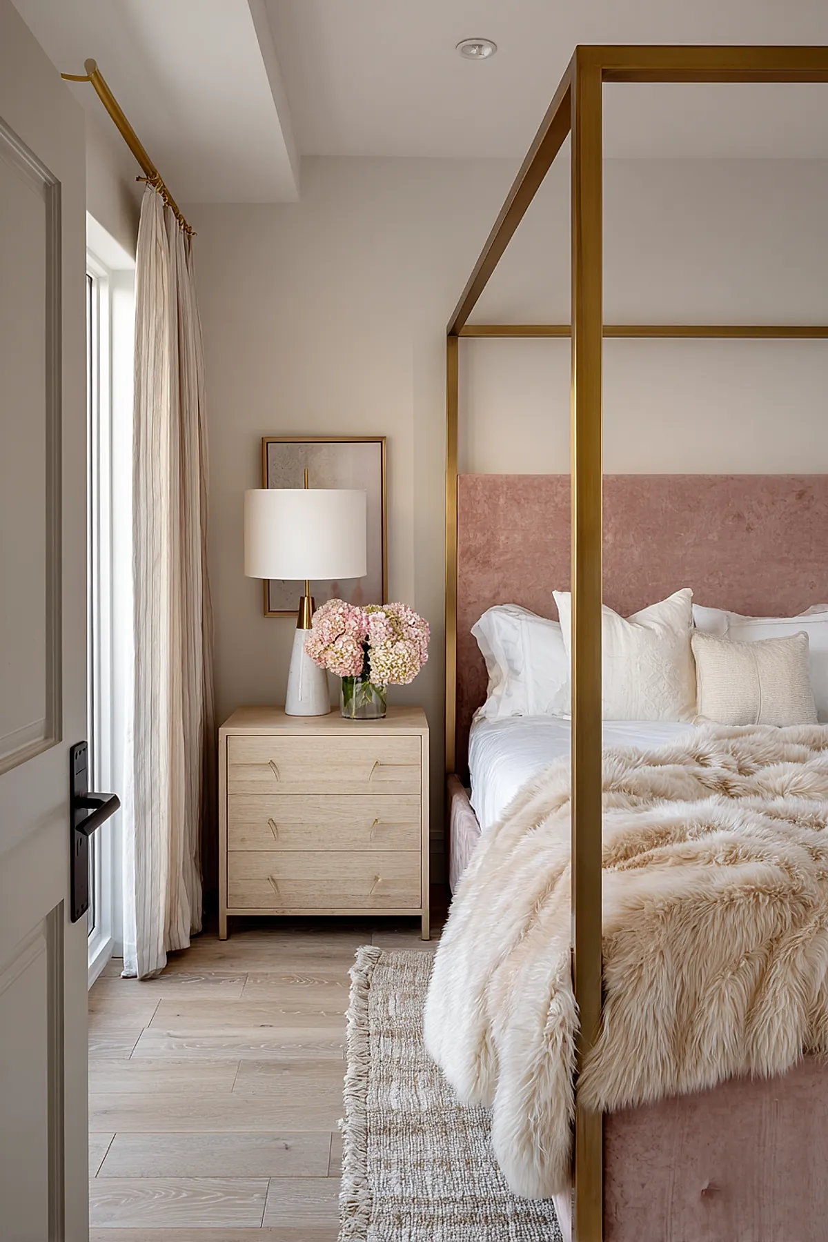 Bedroom with brass metal canopy bed frame, pink velvet upholstered headboard, white bedding with textured pillows, cream faux fur throw, light wood nightstand holding a white lamp and vase of pale pink hydrangeas, neutral walls and curtains.
